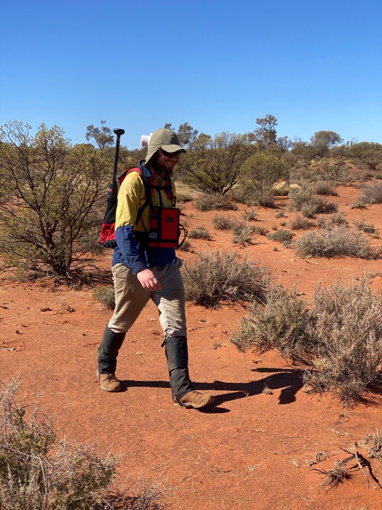Operator in hi-vis walking through red-earth mulga scrub with the compact Modern Mag magnetometer backpack